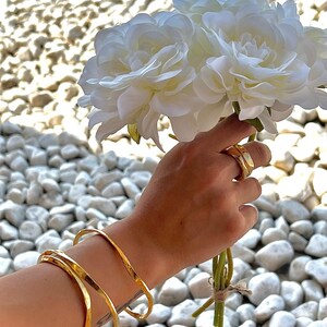 Puede incluir: Una mano sosteniendo un ramo de flores blancas, con brazaletes dorados en la mu&ntilde;eca y anillos dorados en los dedos. Los brazaletes tienen una textura martillada. El fondo es una colecci&oacute;n de piedras blancas.