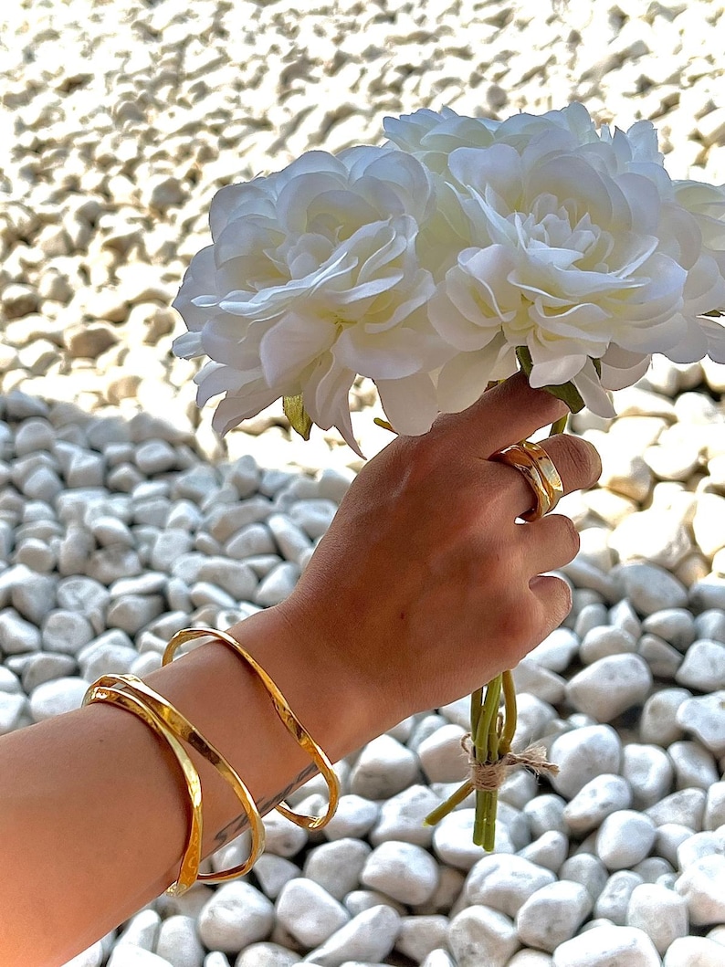 Puede incluir: Una mano sosteniendo un ramo de flores blancas, con brazaletes dorados en la mu&ntilde;eca y anillos dorados en los dedos. Los brazaletes tienen una textura martillada. El fondo es una colecci&oacute;n de piedras blancas.
