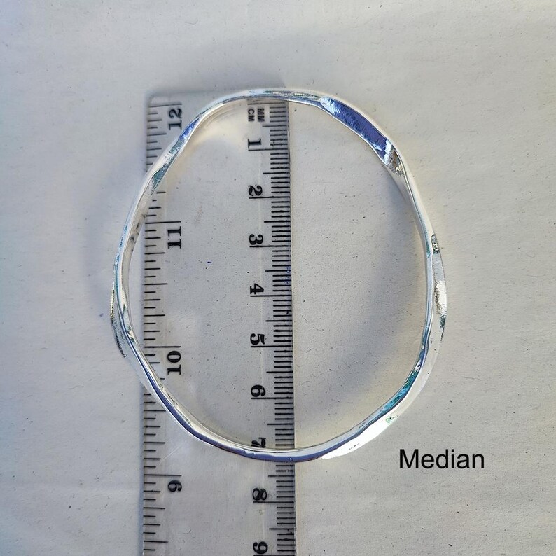 Puede incluir: Un brazalete plateado de estilo martillado. El brazalete tiene aproximadamente 7,6 cm de di&aacute;metro. Se coloca una regla junto al brazalete para medir. La palabra "Median" est&aacute; escrita en la superficie.