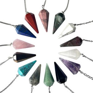 May include: A selection of gemstone pendulums in various colours, including red, blue, pink, and green. Each pendulum is faceted and suspended from a silver chain. The pendulums are arranged in a circular pattern against a white background.