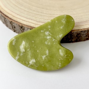 May include: A green jade gua sha tool with white veining. The tool has a smooth, curved shape with a notched edge. It is displayed on a wooden slice, showcasing its natural beauty. This skincare tool is designed for facial massage.