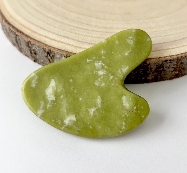 May include: A green jade gua sha tool with white veining. The tool has a smooth, curved shape with a notched edge. It is displayed on a wooden slice, showcasing its natural beauty. This skincare tool is designed for facial massage.