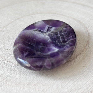 May include: A polished, oval amethyst stone. The stone has a deep purple colour with lighter, crystalline inclusions and patterns. The surface is smooth and reflective, with a slight sheen. The stone is set against a light-coloured, textured wooden surface.