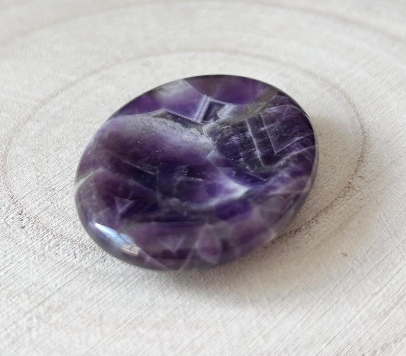 May include: A polished, oval amethyst stone. The stone has a deep purple colour with lighter, crystalline inclusions and patterns. The surface is smooth and reflective, with a slight sheen. The stone is set against a light-coloured, textured wooden surface.