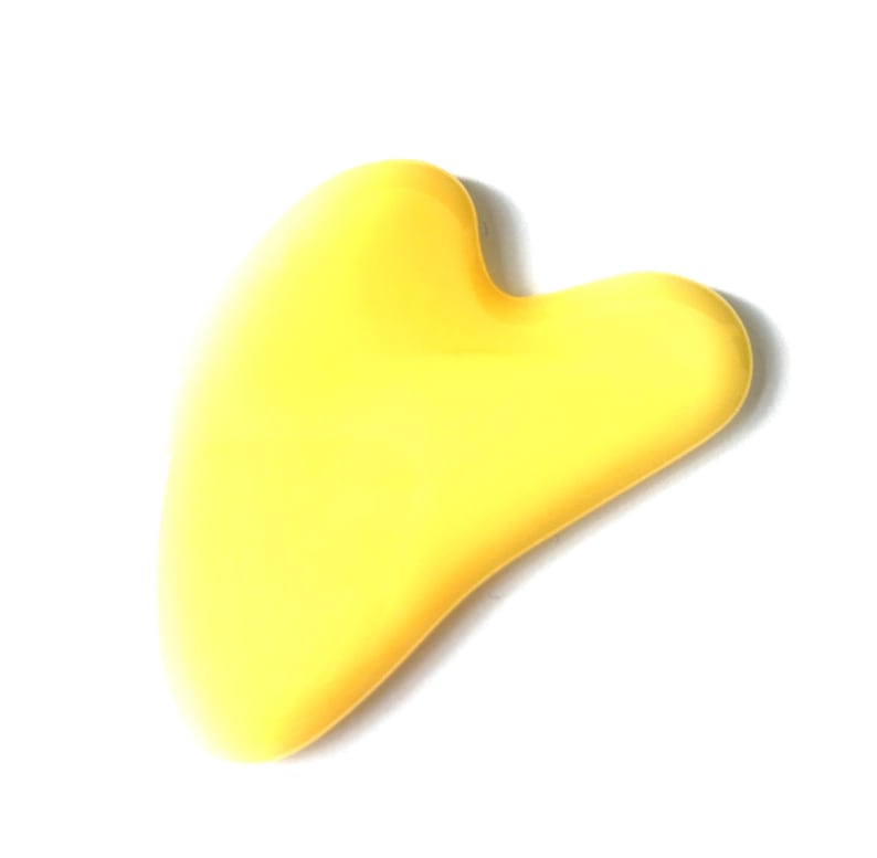 May include: A yellow gua sha tool in a heart shape, designed for facial massage. The smooth, curved edges are visible against a white background. This skincare tool is made of a solid, non-porous material.