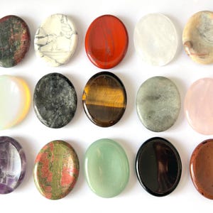 May include: A collection of fifteen oval gemstones in diverse colours and patterns. The stones feature red, white, black, yellow, green, and pink hues. The gemstones are arranged in three rows of five, displaying their unique textures and colours.