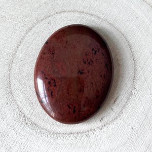 May include: A polished, oval-shaped stone in a deep burgundy hue with black inclusions. The stone has a glossy finish and reflects light. The overall shape is smooth and rounded, suggesting it is a palm stone or worry stone.