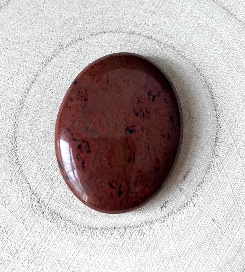 May include: A polished, oval-shaped stone in a deep burgundy hue with black inclusions. The stone has a glossy finish and reflects light. The overall shape is smooth and rounded, suggesting it is a palm stone or worry stone.