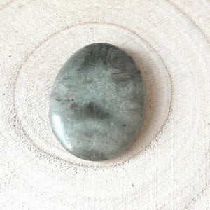 May include: Oval-shaped, polished gemstone with a mix of grey and pale green hues. The stone has a smooth, glossy surface and subtle dark grey veining. The gemstone is set against a light-coloured, textured wooden surface.