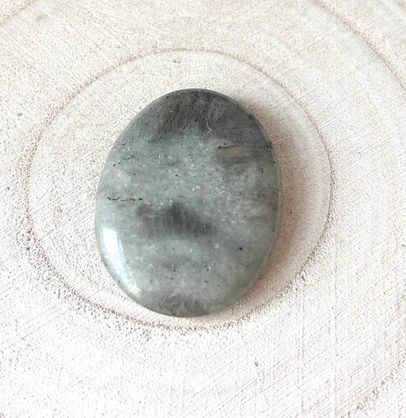 May include: Oval-shaped, polished gemstone with a mix of grey and pale green hues. The stone has a smooth, glossy surface and subtle dark grey veining. The gemstone is set against a light-coloured, textured wooden surface.