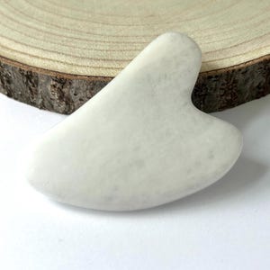 May include: A white, heart-shaped gua sha tool made of stone. The tool has a smooth, polished surface and is designed for facial massage. The gua sha is displayed on a wooden slice with a white background.