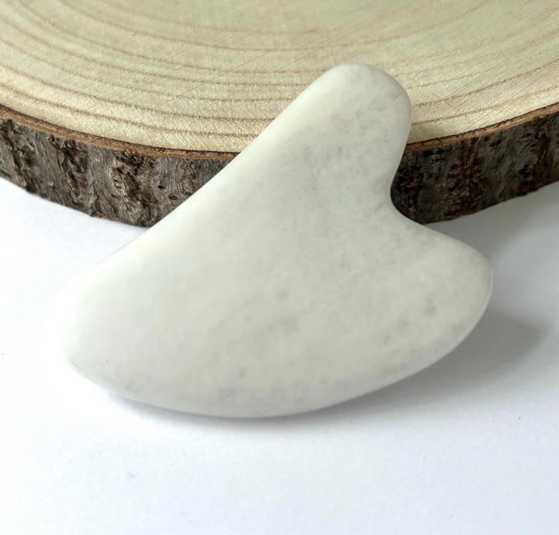 May include: A white, heart-shaped gua sha tool made of stone. The tool has a smooth, polished surface and is designed for facial massage. The gua sha is displayed on a wooden slice with a white background.