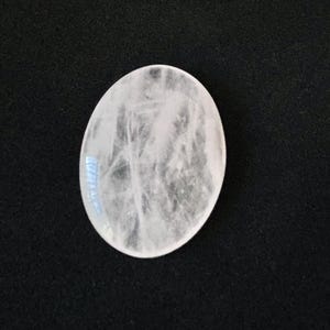 May include: A pale, oval-shaped gemstone with a cloudy internal texture. The stone is a milky white colour and appears polished. The background is a solid black, providing contrast to the light-coloured stone.