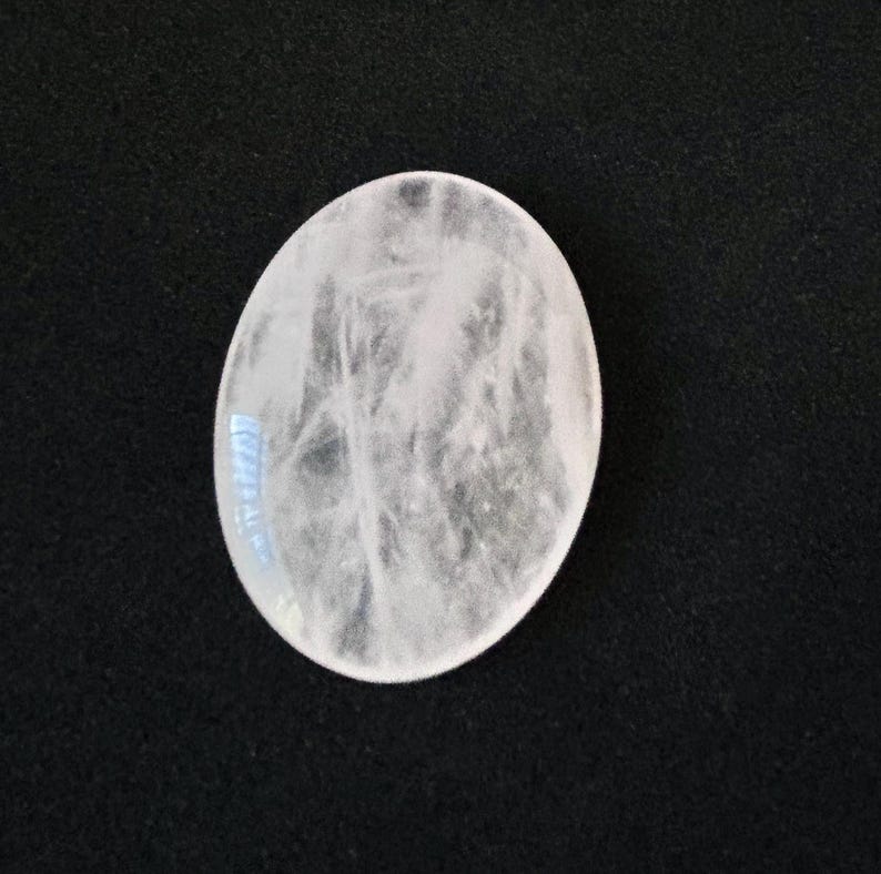 May include: A pale, oval-shaped gemstone with a cloudy internal texture. The stone is a milky white colour and appears polished. The background is a solid black, providing contrast to the light-coloured stone.