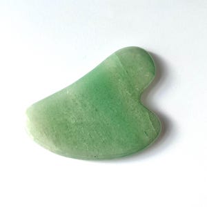 May include: A jade green Gua Sha tool with a smooth, curved design. The tool is made of a translucent, light green stone and is designed for facial massage and skincare routines.