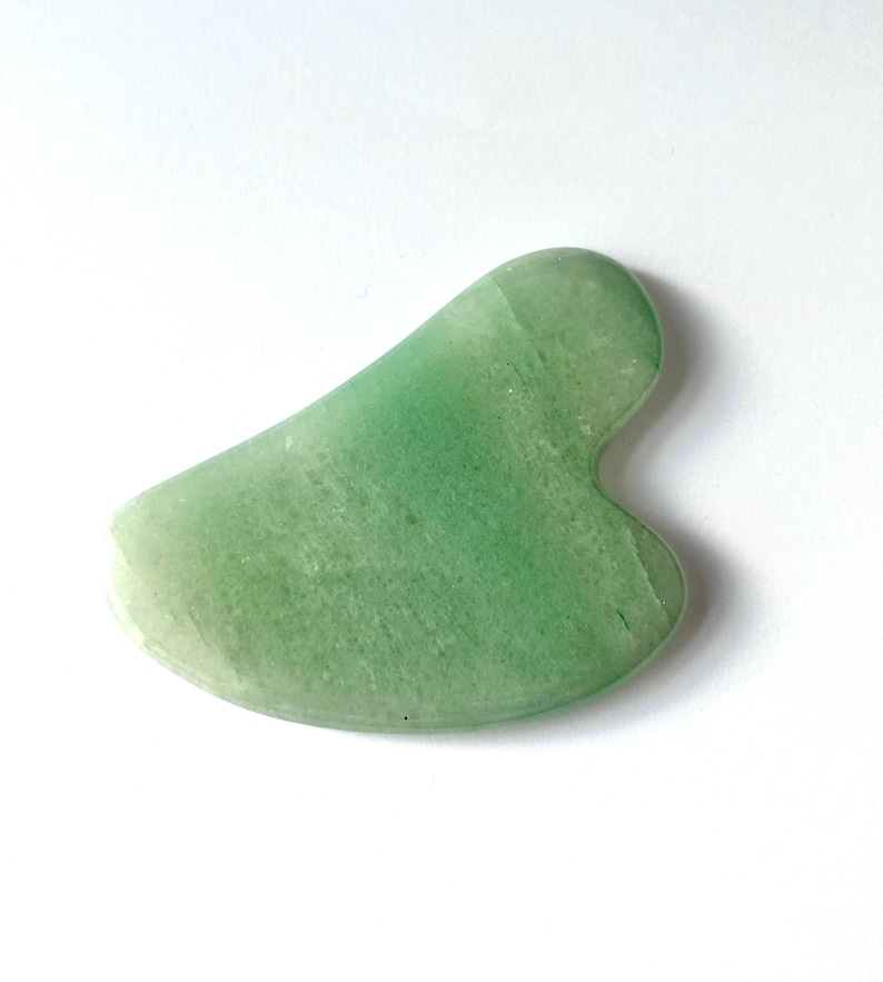 May include: A jade green Gua Sha tool with a smooth, curved design. The tool is made of a translucent, light green stone and is designed for facial massage and skincare routines.