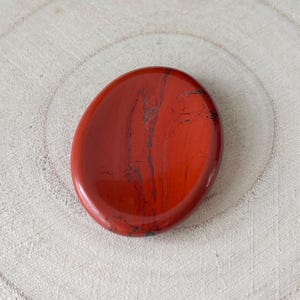 May include: A polished, oval red jasper stone with dark grey veining. The stone has a smooth, glossy finish and a slightly concave surface. The jasper is set against a light-coloured, textured wooden background.