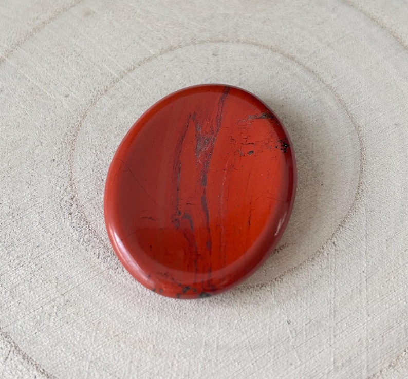 May include: A polished, oval red jasper stone with dark grey veining. The stone has a smooth, glossy finish and a slightly concave surface. The jasper is set against a light-coloured, textured wooden background.