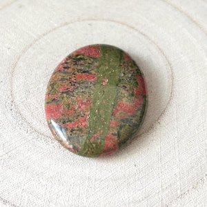 May include: Polished oval unakite stone. The stone displays a blend of green, red, and black hues, with a distinct green stripe through the middle. The stone is set against a light-coloured, textured background, creating a natural aesthetic.