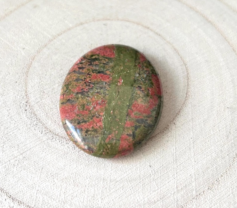 May include: Polished oval unakite stone. The stone displays a blend of green, red, and black hues, with a distinct green stripe through the middle. The stone is set against a light-coloured, textured background, creating a natural aesthetic.