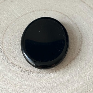 May include: A glossy, oval black stone. The stone has a smooth, reflective surface and is set against a light-coloured, textured background. The stone's shape is slightly elongated, and it appears to be a natural gemstone.