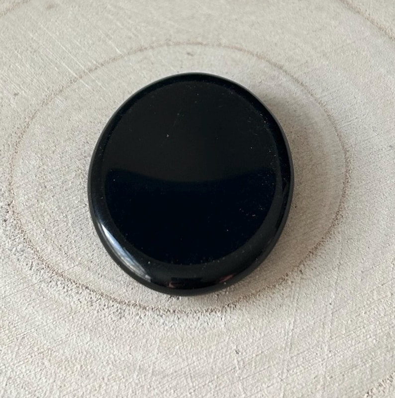 May include: A glossy, oval black stone. The stone has a smooth, reflective surface and is set against a light-coloured, textured background. The stone's shape is slightly elongated, and it appears to be a natural gemstone.