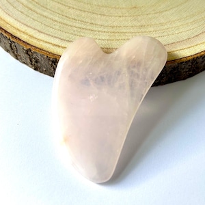 May include: A light pink rose quartz gua sha tool with a curved, heart-like shape. The smooth stone is displayed on a light wood slice with a white background. This facial massage tool is used for skincare.