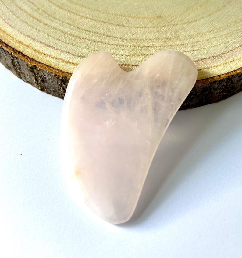May include: A light pink rose quartz gua sha tool with a curved, heart-like shape. The smooth stone is displayed on a light wood slice with a white background. This facial massage tool is used for skincare.