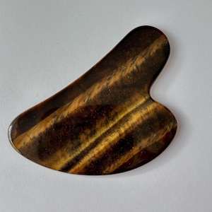 May include: A tiger's eye gua sha tool. The stone is a mix of brown and gold with a smooth, polished surface. The tool has a curved shape with a rounded end and a notched edge.