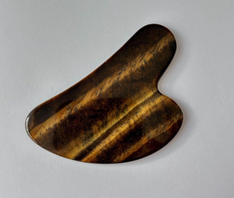 May include: A tiger's eye gua sha tool. The stone is a mix of brown and gold with a smooth, polished surface. The tool has a curved shape with a rounded end and a notched edge.