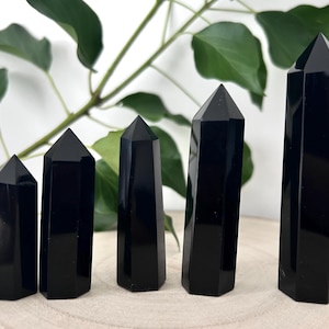 May include: Five polished black obsidian crystal towers arranged in a row from shortest to tallest. Each tower features a smooth, reflective surface with a hexagonal base and a pointed, faceted top, showcasing a deep, solid black colour throughout.