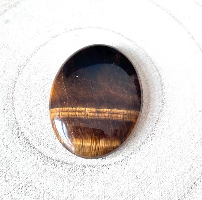 May include: Polished oval tiger's eye gemstone. The stone displays bands of brown, gold, and black, forming a striped pattern. The gemstone has a smooth, reflective surface and a natural, earthy aesthetic. The stone is approximately 4 cm in length.