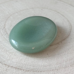May include: A polished, oval-shaped, pale green gemstone, possibly aventurine, sits on a light-coloured, textured wooden surface. The stone has a smooth, reflective surface, with slight colour and texture variations. The gemstone is a natural, organic item.