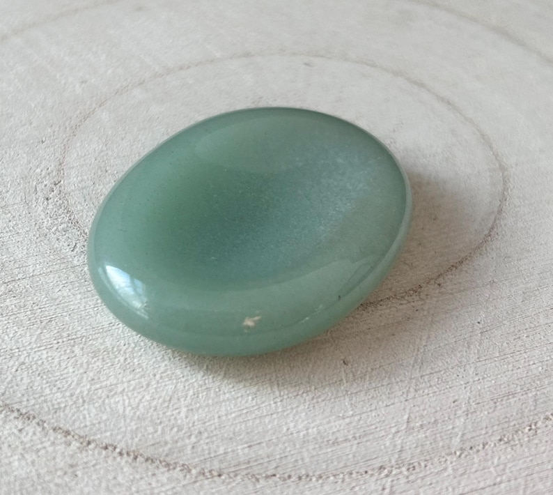 May include: A polished, oval-shaped, pale green gemstone, possibly aventurine, sits on a light-coloured, textured wooden surface. The stone has a smooth, reflective surface, with slight colour and texture variations. The gemstone is a natural, organic item.