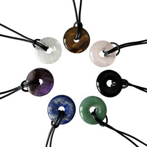 May include: A selection of gemstone donut pendants on black cords. The pendants are in various colours, including white, brown, pink, purple, blue, black, and green. Each pendant has a hole in the centre.