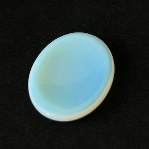 May include: A light blue, oval-shaped stone with a white border. The stone has a smooth, polished surface and is set against a black background. The stone is a pale, pastel colour.