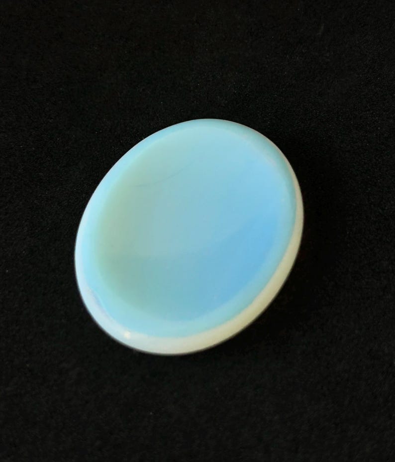 May include: A light blue, oval-shaped stone with a white border. The stone has a smooth, polished surface and is set against a black background. The stone is a pale, pastel colour.