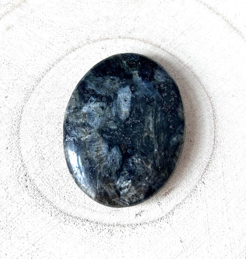 May include: Oval-shaped, polished stone with a dark blue and grey marbled pattern. The stone has a smooth, glossy surface and is set against a white, textured background. The stone's colour variations create a natural, organic aesthetic.
