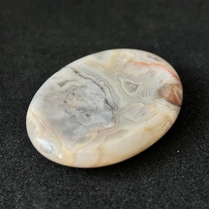 May include: An oval polished stone displaying a swirling pattern of grey, cream, and reddish-brown tones. The stone has a smooth, glossy surface, set against a dark backdrop. The natural patterns create a unique, organic aesthetic, suitable for jewellery.