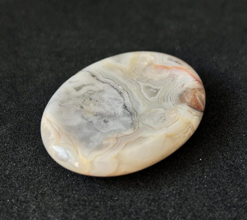 May include: An oval polished stone displaying a swirling pattern of grey, cream, and reddish-brown tones. The stone has a smooth, glossy surface, set against a dark backdrop. The natural patterns create a unique, organic aesthetic, suitable for jewellery.