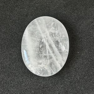 May include: A clear, oval-shaped quartz crystal. The stone is translucent with internal veining and sits on a dark grey surface. The crystal is smooth and polished, reflecting light.