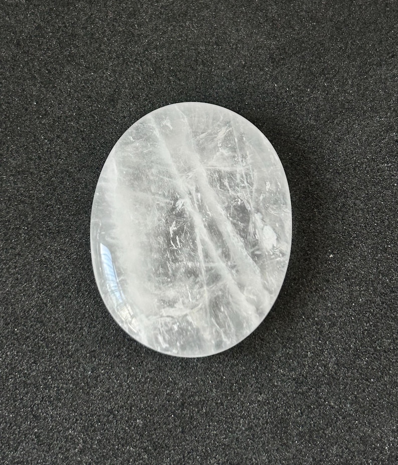 May include: A clear, oval-shaped quartz crystal. The stone is translucent with internal veining and sits on a dark grey surface. The crystal is smooth and polished, reflecting light.