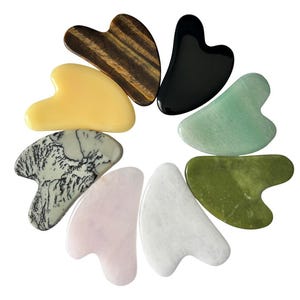May include: A collection of eight heart-shaped gua sha tools in various colors and materials. The tools include a tiger's eye, black obsidian, jade, and rose quartz. The arrangement is circular, showcasing the diverse textures and hues.