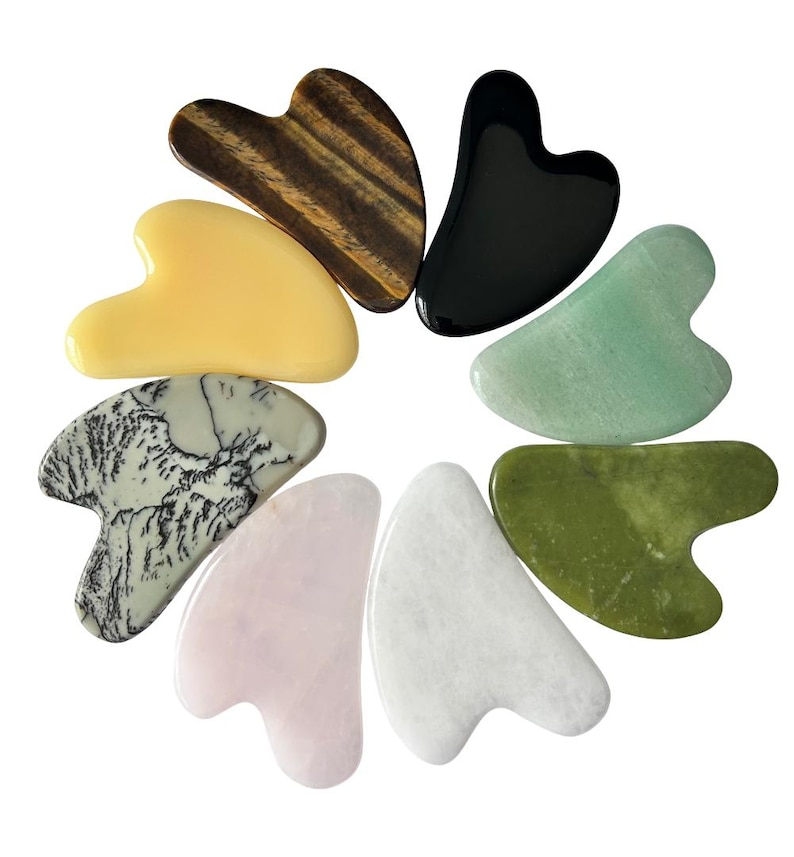 May include: A collection of eight heart-shaped gua sha tools in various colors and materials. The tools include a tiger's eye, black obsidian, jade, and rose quartz. The arrangement is circular, showcasing the diverse textures and hues.