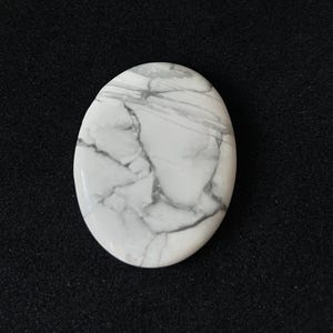 May include: Oval-shaped white gemstone with grey veining. The polished stone has a smooth surface and a marbled appearance. The stone is set against a black background.