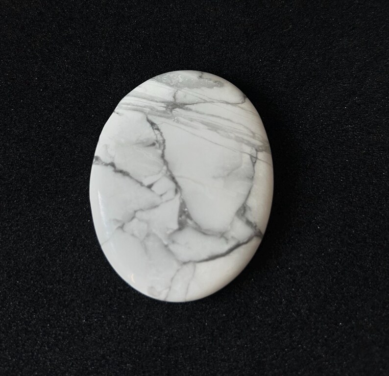 May include: Oval-shaped white gemstone with grey veining. The polished stone has a smooth surface and a marbled appearance. The stone is set against a black background.