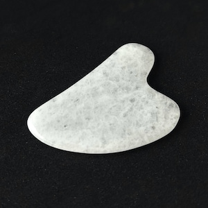 May include: A white and gray gua sha tool with a heart-shaped notch. The smooth, polished stone has a marbled appearance and is designed for facial massage. The tool is set against a black background.