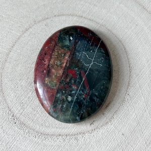 May include: Oval gemstone with a polished surface. The stone displays a mix of colours, including deep red, green, and dark grey, with white veining. The gemstone is presented on a light-coloured, textured wooden surface.