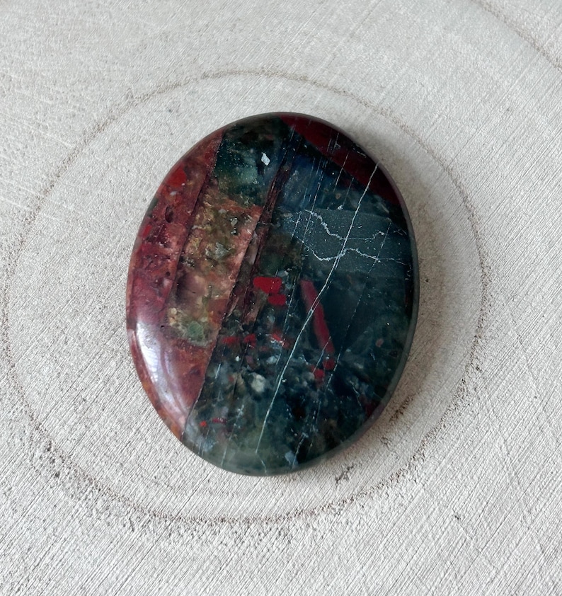 May include: Oval gemstone with a polished surface. The stone displays a mix of colours, including deep red, green, and dark grey, with white veining. The gemstone is presented on a light-coloured, textured wooden surface.