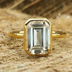 May include: A gold ring featuring an emerald-cut diamond. The diamond is set in a bezel setting and has a clear, brilliant finish.